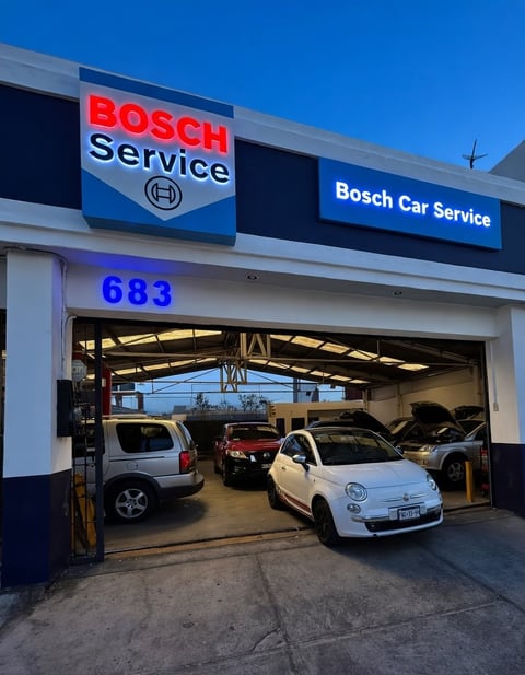 Bosch Car Service garage exterior with number 683, showing white and dark vehicles inside the open bay doors against a clear blue sky